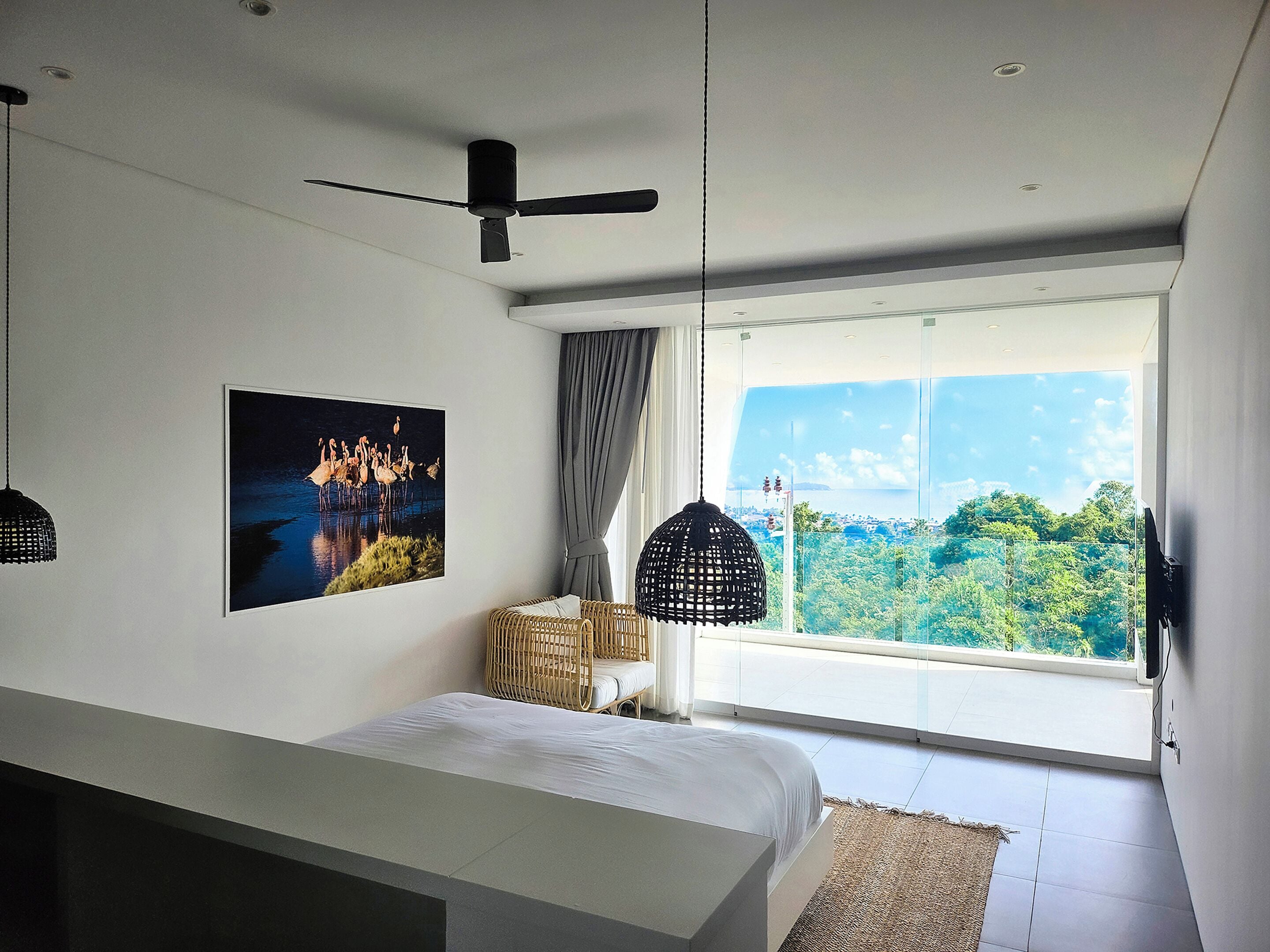 Een slaapkamer met een bed, een plafondventilator, een groot raam met uitzicht op groen en een ingelijste foto aan de muur.