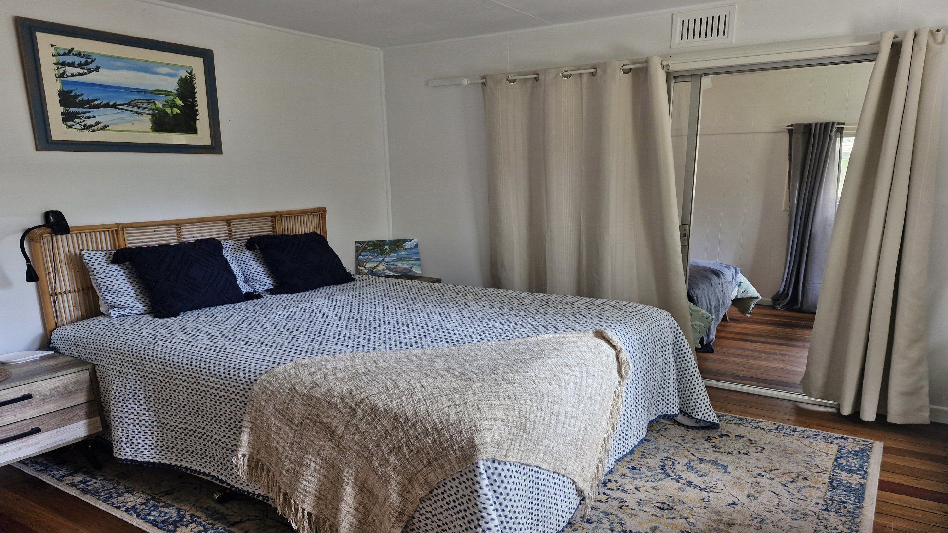 A bedroom with a bed, a nightstand, a lamp, a painting, a window with curtains, and a wooden floor.
