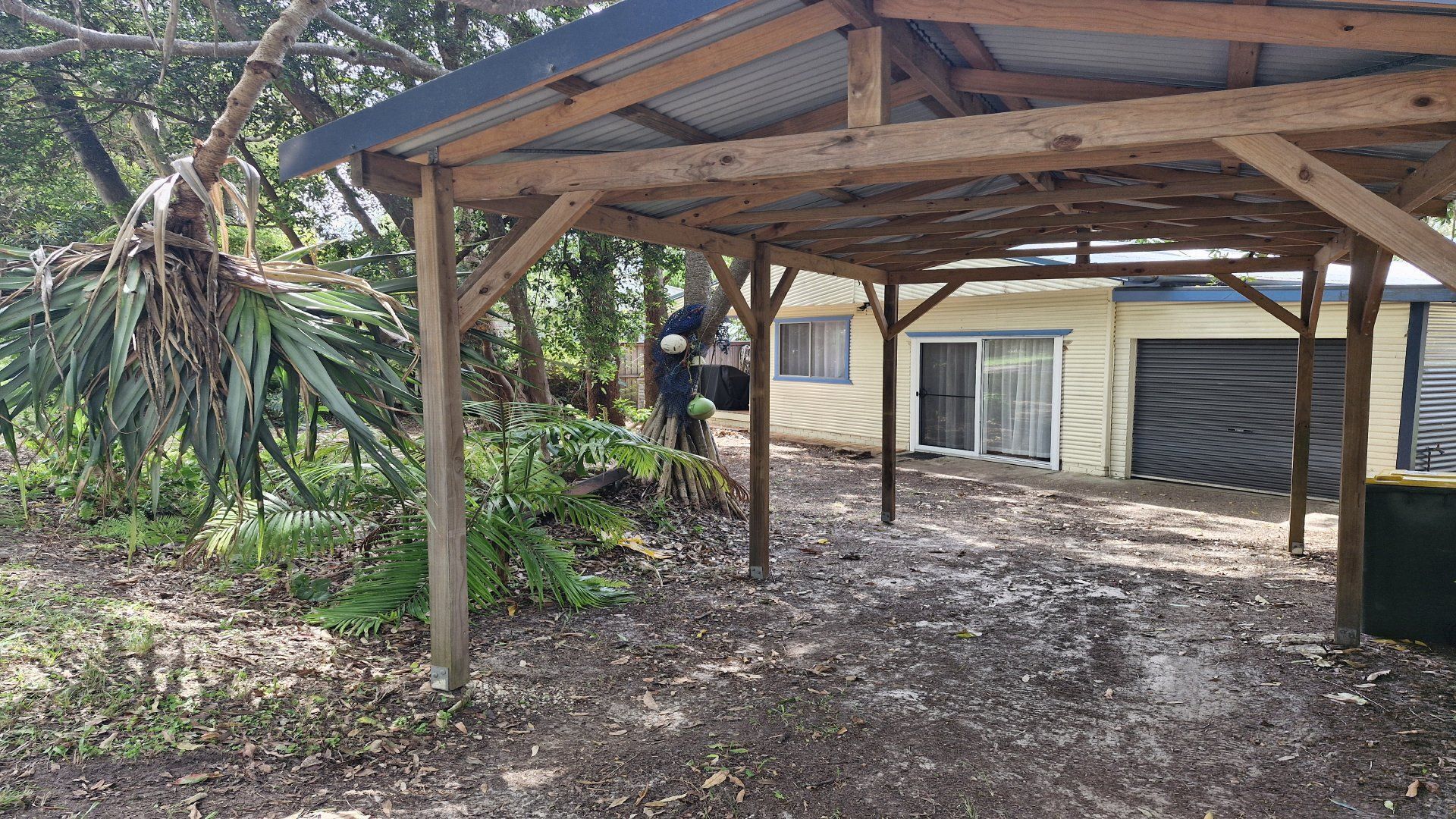 A covered outdoor area with a wooden structure, a dirt ground, and a building in the background.