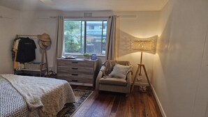 Quarto com cama, penteadeira, cadeira, abajur e janela vista para área verde.