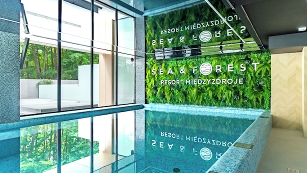 Uma área moderna de piscina interna com paredes de vidro, vista para vegetação externa e o texto "SEA & FOREST RESORT MIEDZYDROJE".