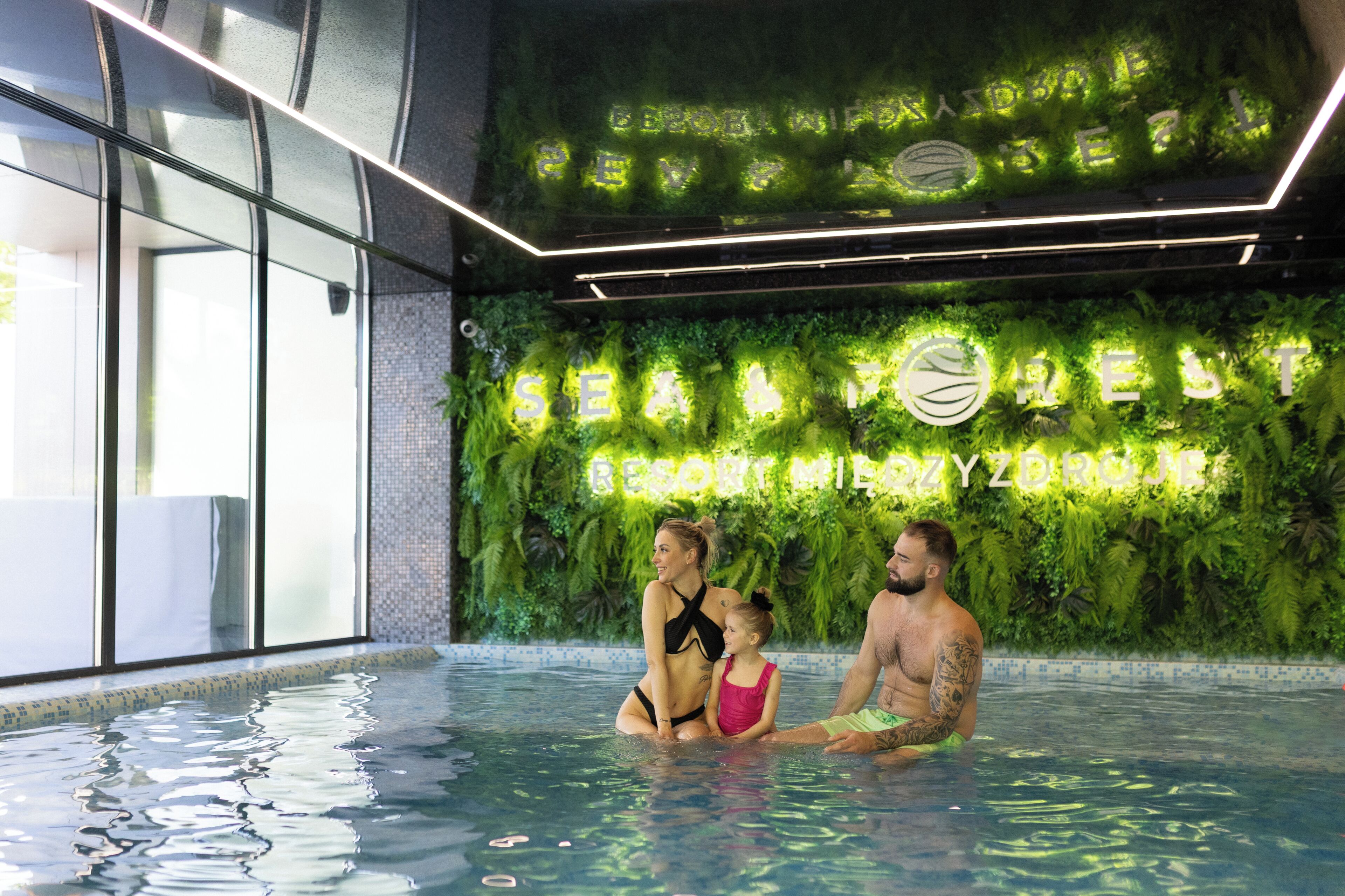 Une piscine où une famille se détend, entourée de plantes vertes à l’intérieur, et un panneau néon indiquant « SEA RESORT ».