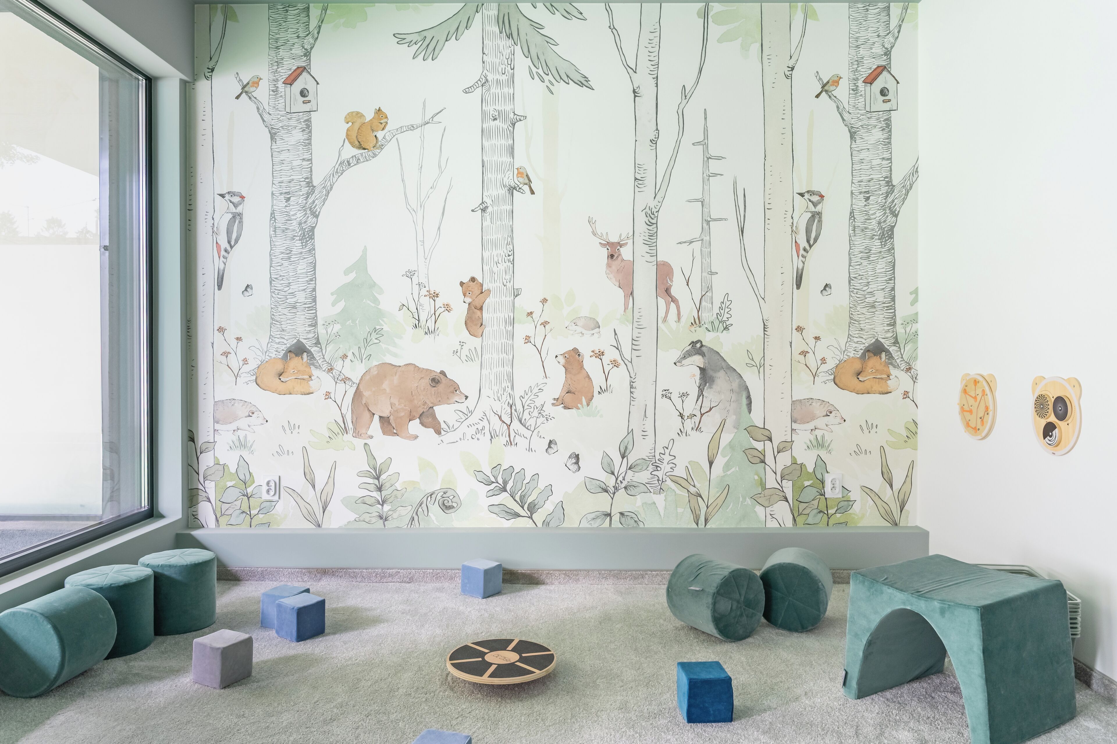 Ein Zimmer mit rauenholzinspirierter Tapete, einem Fenster und einer Spielgegend mit Kissen und Bauklötzen.