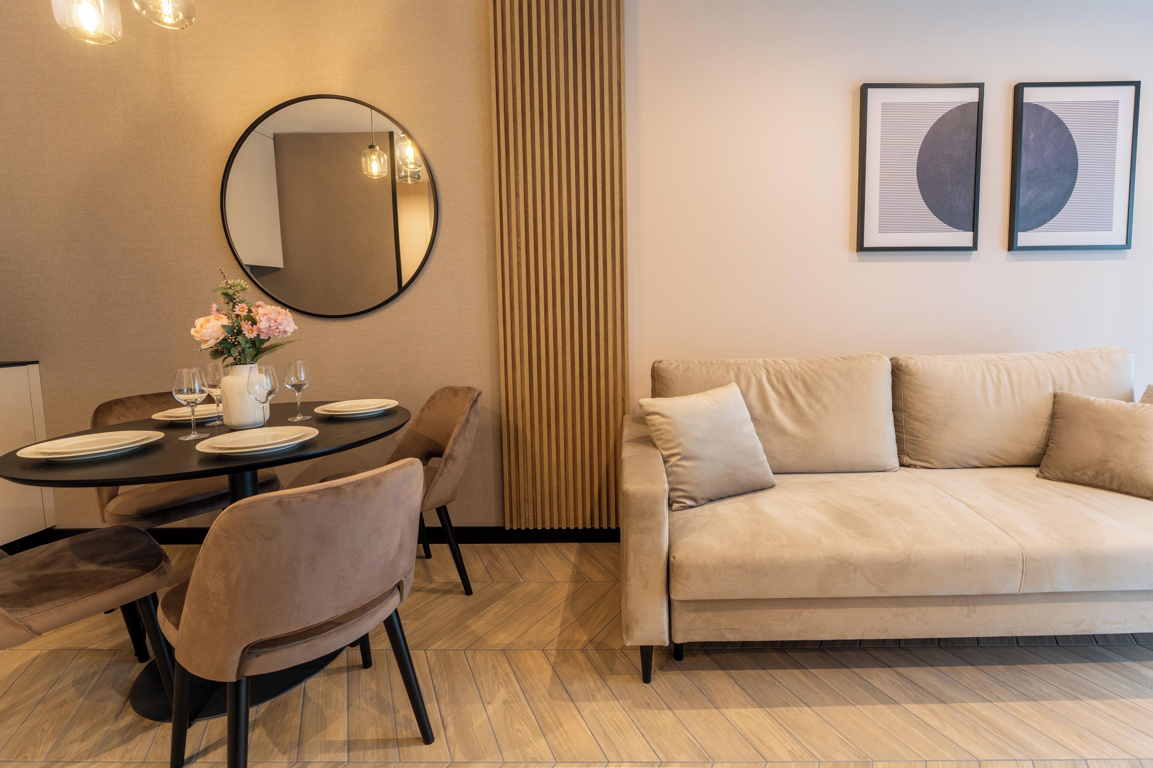 Un salon moderne avec un canapé beige, une table de salle à manger ronde entourée de chaises, un grand miroir et deux œuvres d’art encadrées accrochées au mur.