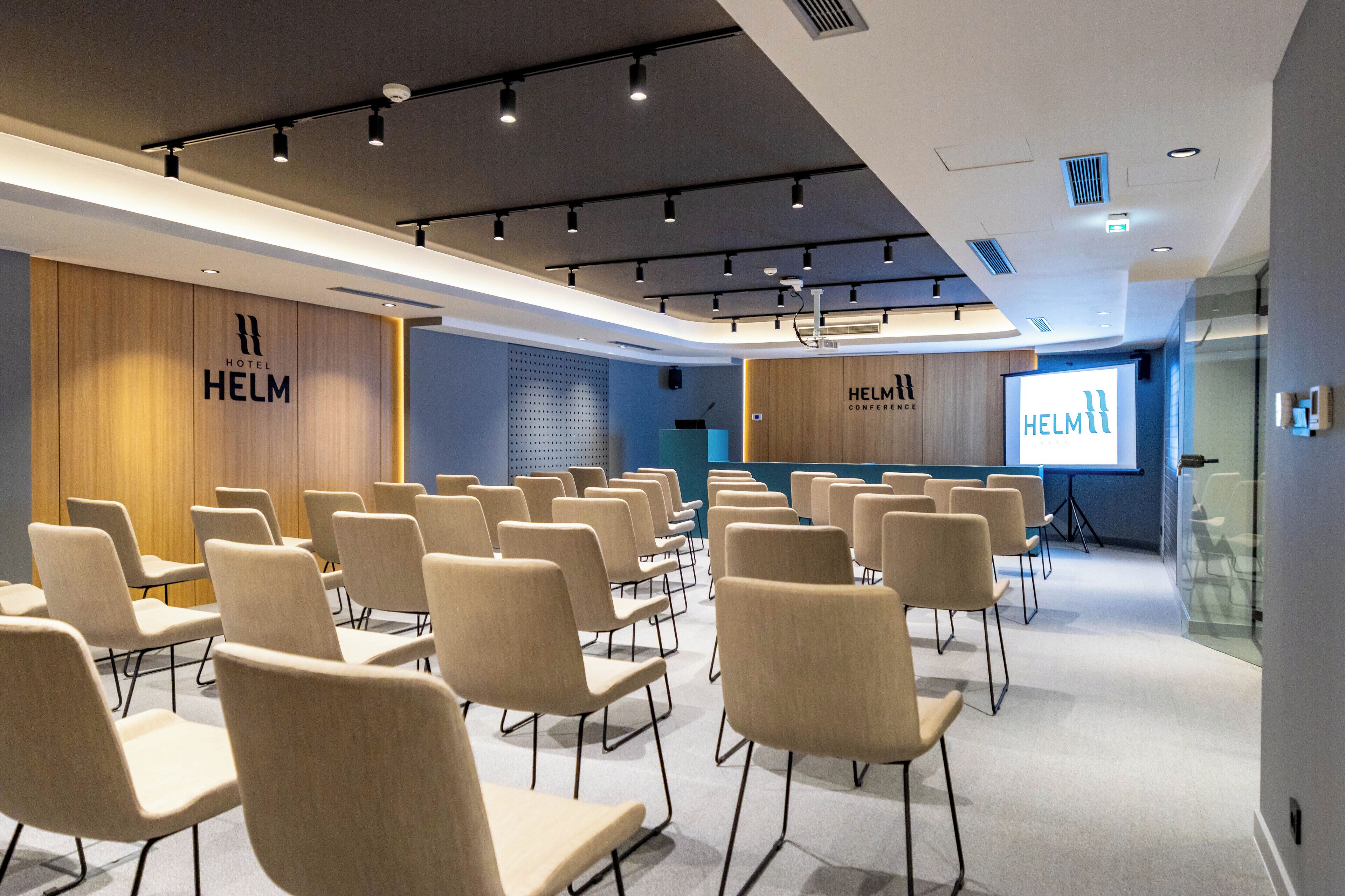 Une salle de conférence avec des rangées de chaises beiges, un pupitre et un écran de projection affichant le nom de l’hôtel.