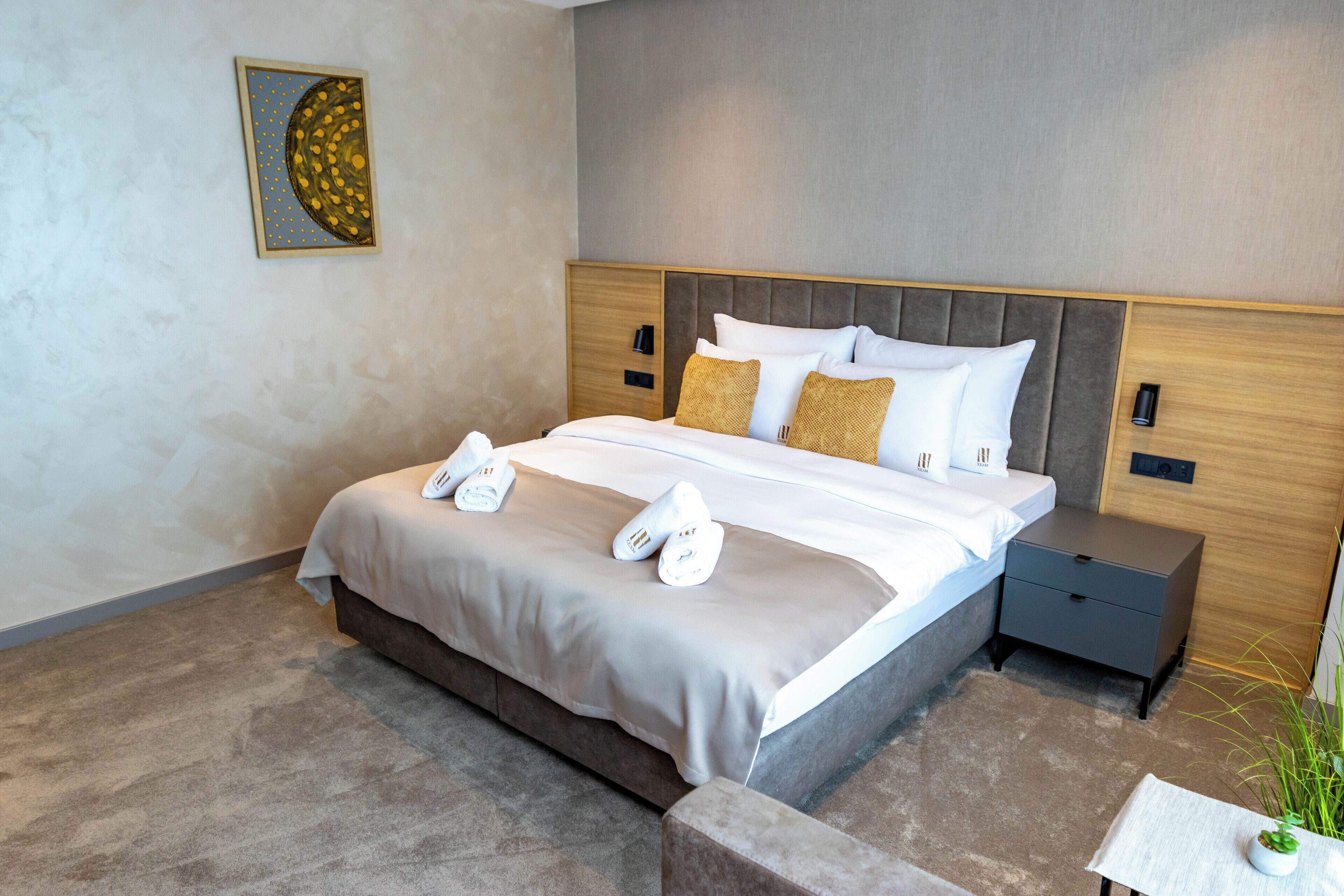 A modern hotel room with a large bed, a nightstand, a small table with a plant, and a wall art piece.