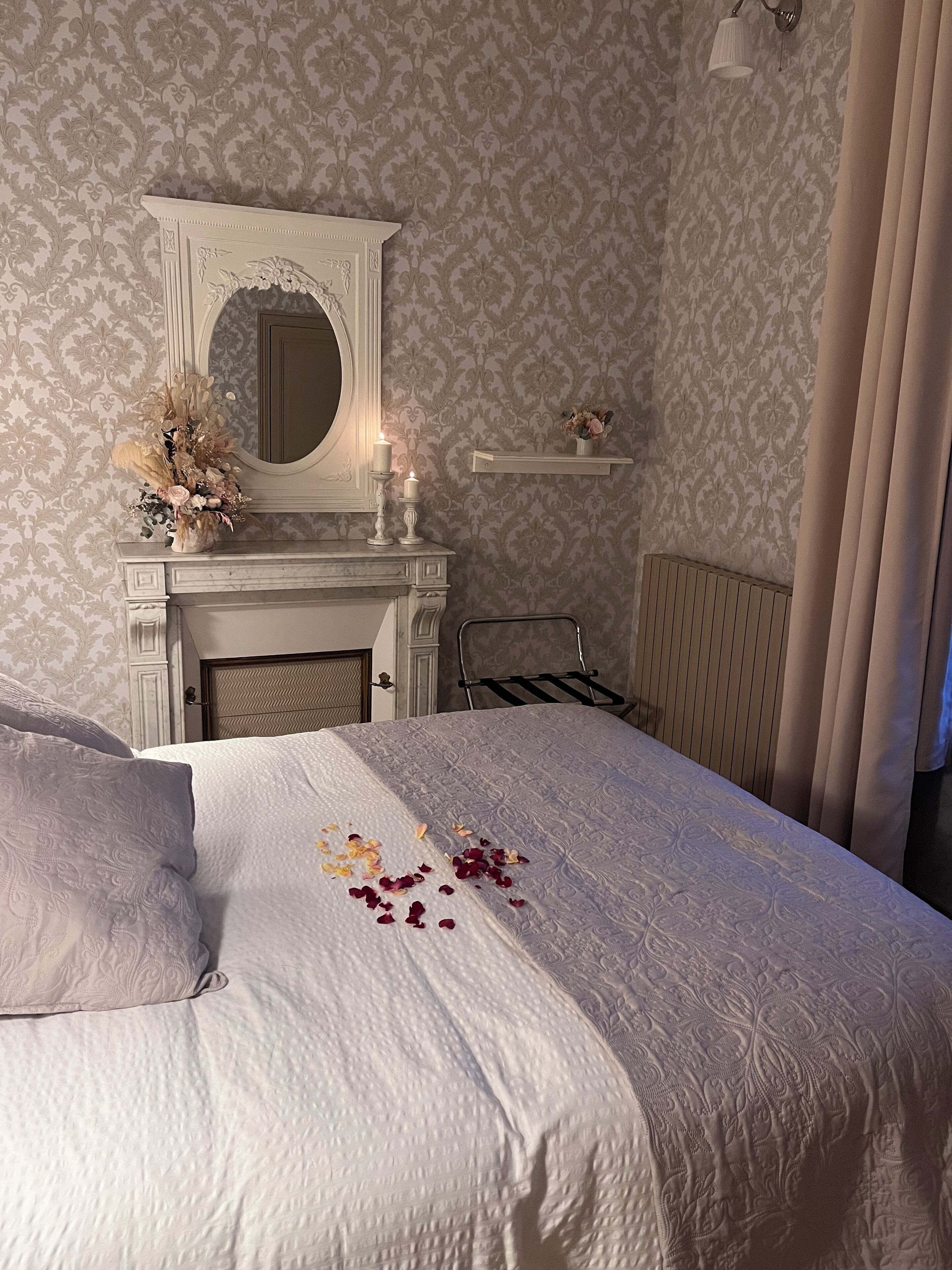 Un dormitorio con una cama grande, una chimenea, un espejo y una estantería con flores.
