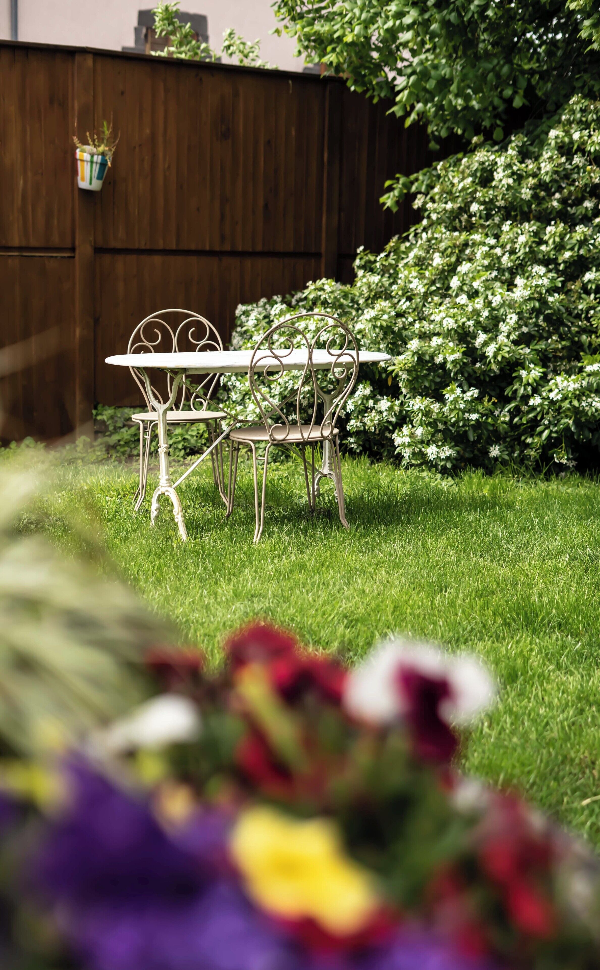 A garden patio with a table and chairs, surrounded by lush greenery and flowering plants.