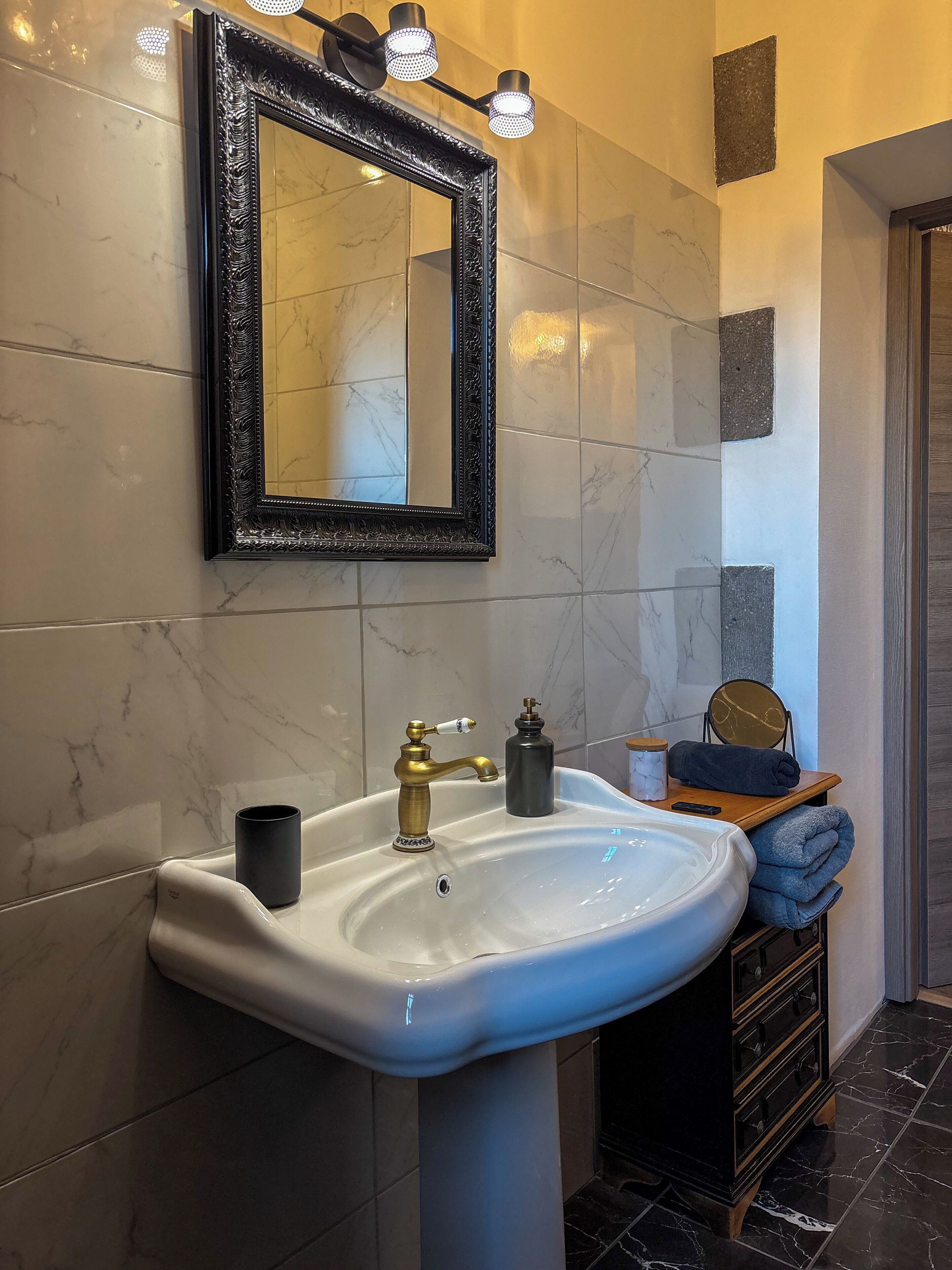 A bathroom with a white sink, gold faucet, and a black mirror with lights. There is a wooden shelf with towels and a black bottle.