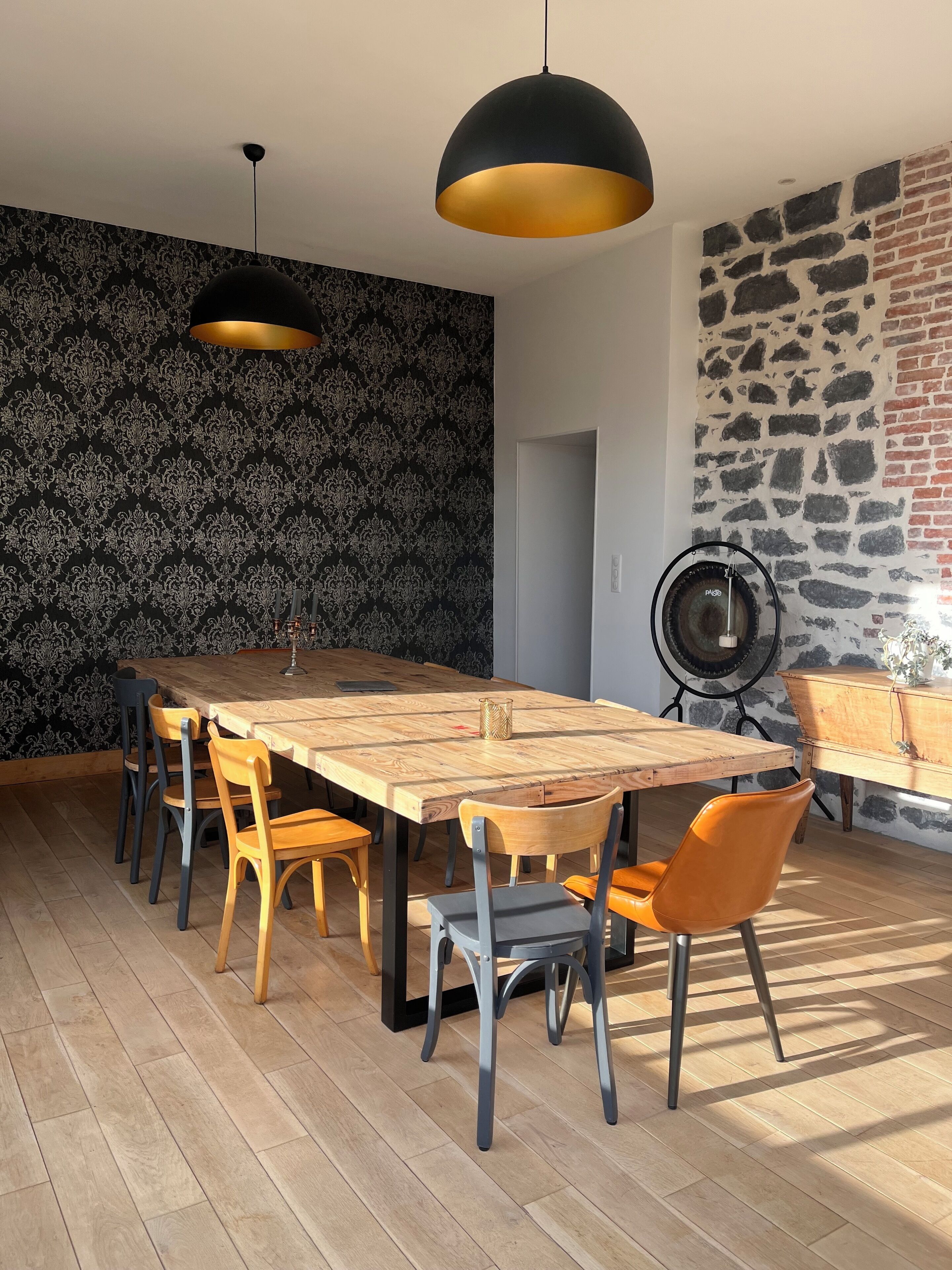 Ein Essbereich mit einem Holztisch, Stühlen, einer schwarzen und goldenen Pendelleuchte sowie einer Wand mit Muster.