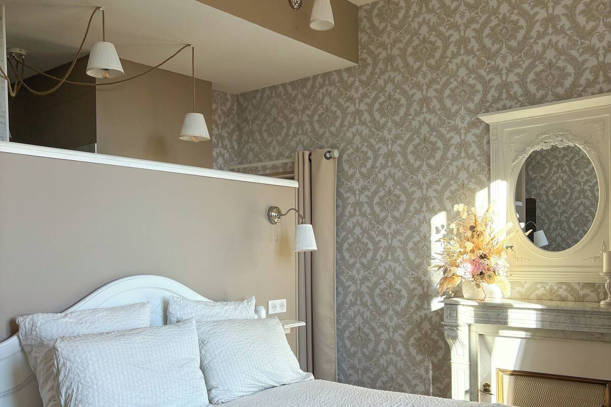 A neatly arranged bedroom with a bed, bedside lamps, a mirror, and a fireplace.