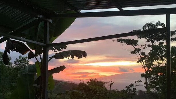 Blick von einer überdachten Außenfläche mit Metalldach auf eine Landschaft mit Hügeln und Sonnenuntergang.