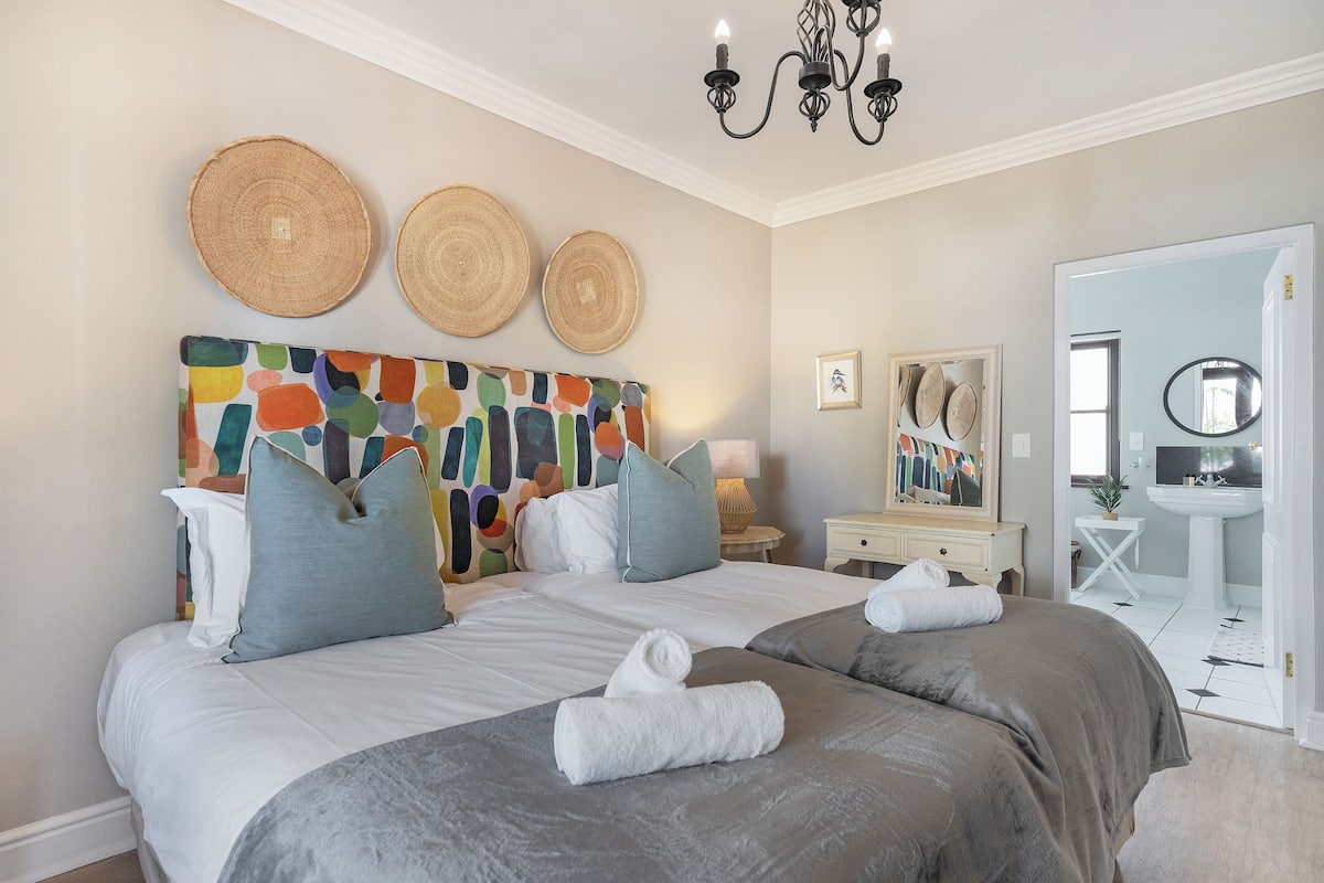 A bedroom with two beds, a colorful abstract headboard, and a bathroom visible through an open door.