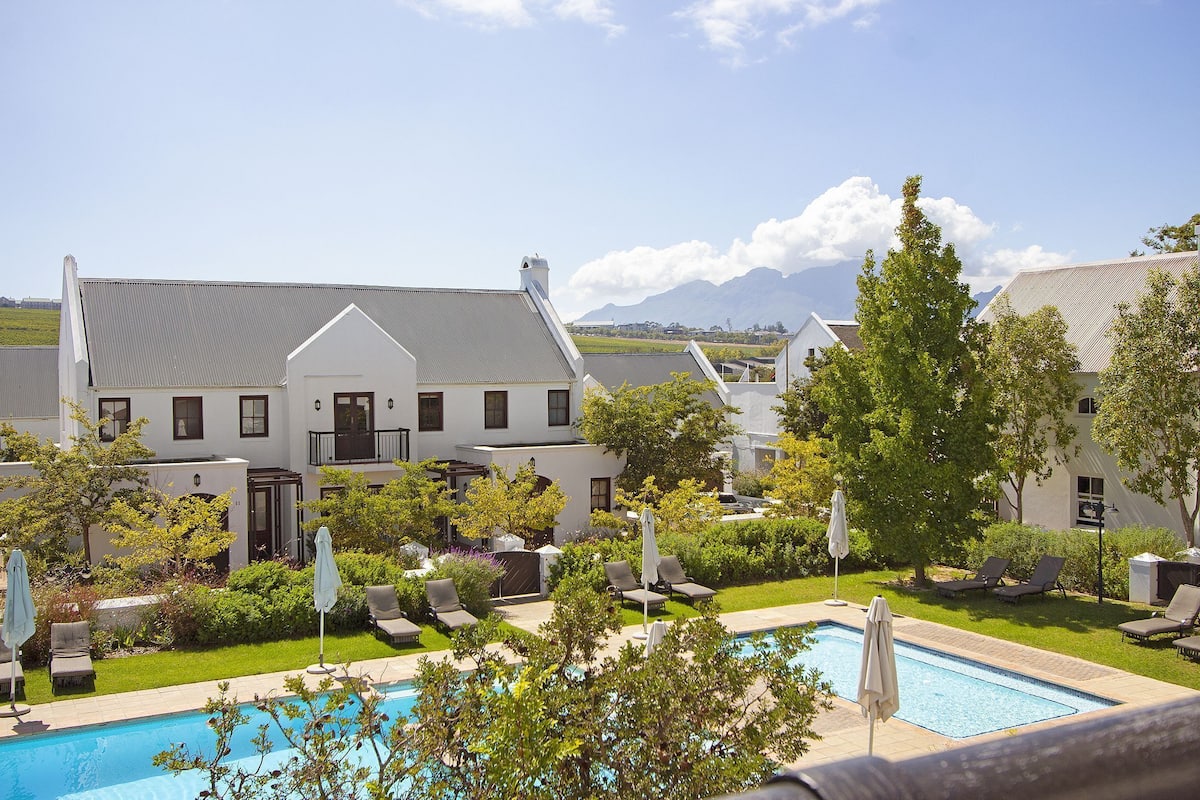 A pool area with lounge chairs and umbrellas, surrounded by greenery and a large building with a sloped roof.