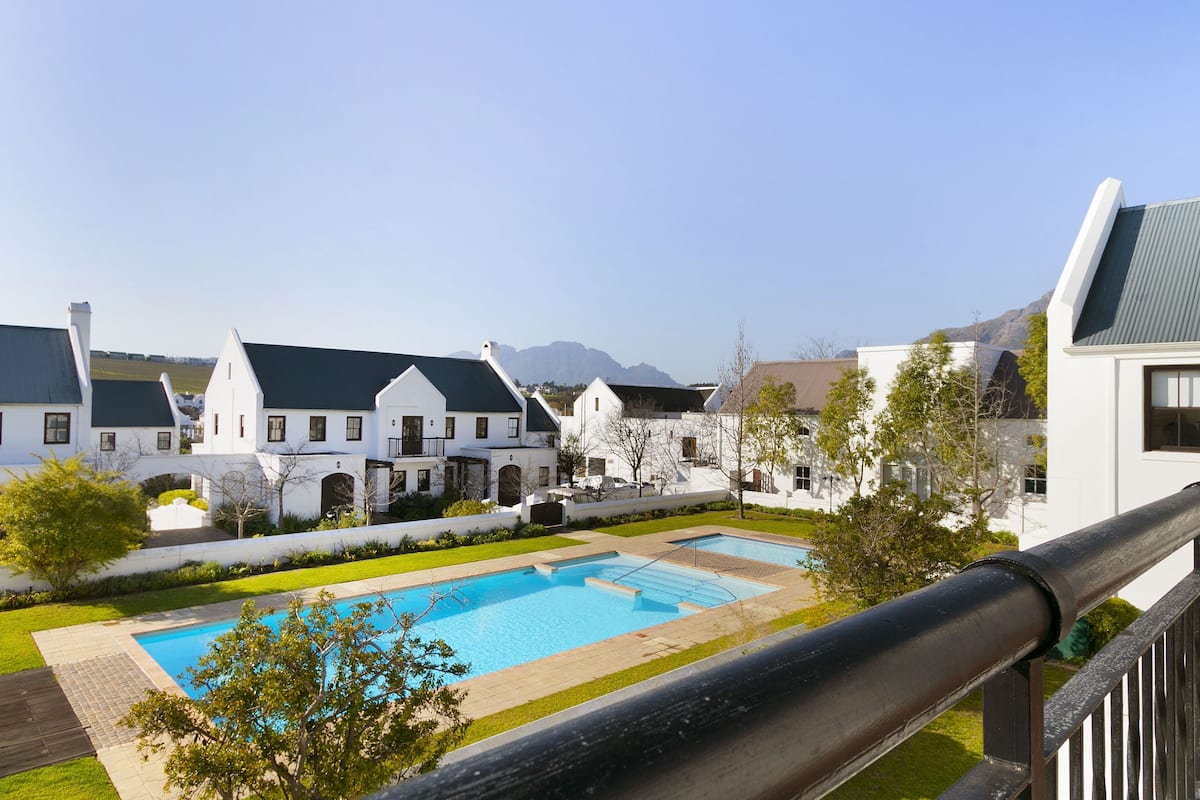 A swimming pool surrounded by white buildings with green roofs, set against a mountainous backdrop.