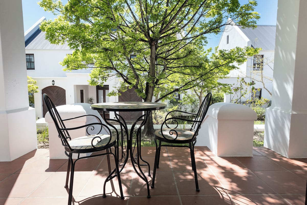 A patio with a round table and two chairs, a tree, and a white building in the background.