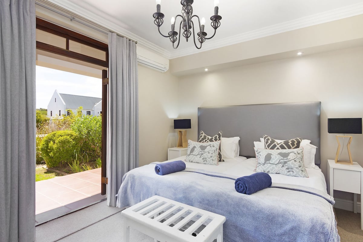 A bedroom with a large bed, two bedside tables, a chandelier, and a view of greenery outside.