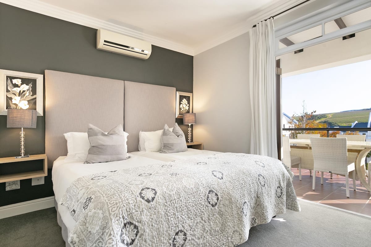 A bedroom with a large bed, two bedside tables with lamps, a view of the outdoors through a sliding door, and an air conditioning unit on the wall.