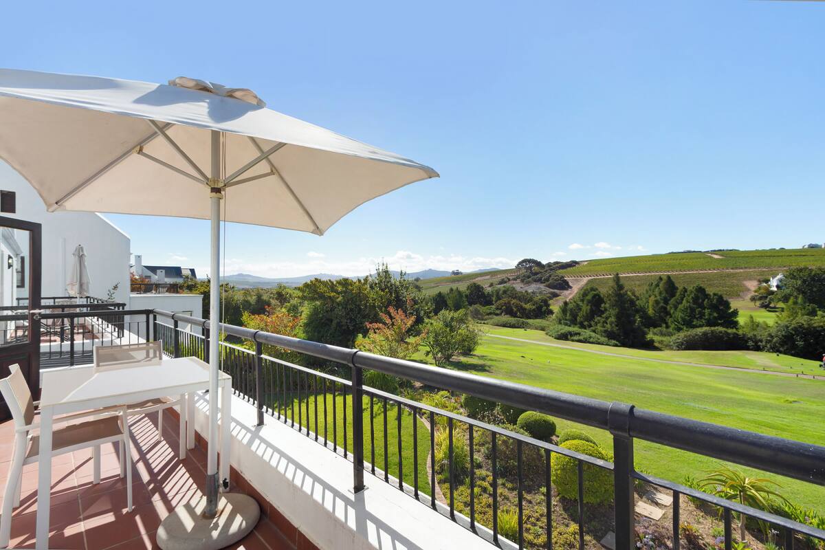 A balcony with a white table and chair, a large umbrella, and a view of a green landscape.
