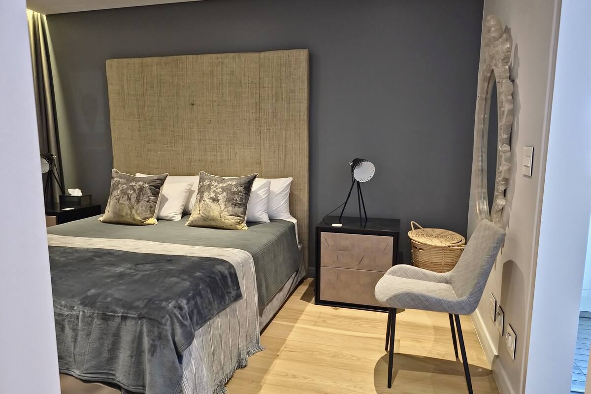 A bedroom with a large bed, a bedside table with a lamp, a chair, and a wall-mounted mirror.