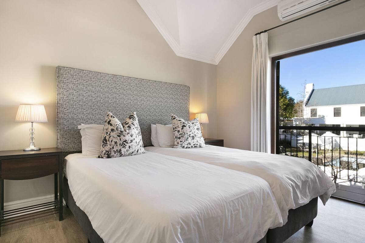 A hotel room with a large bed, two bedside tables with lamps, a patterned headboard, and a view of a balcony with outdoor furniture.
