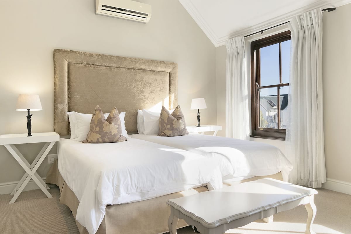 A hotel room with two beds, a large headboard, a window with curtains, and a small table.