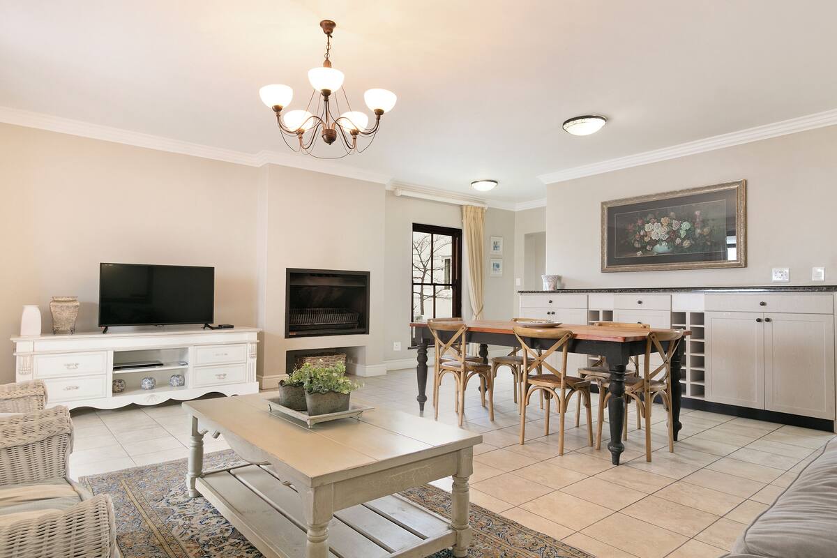 A living room with a TV, a fireplace, a dining area with chairs, and a kitchen with cabinets.