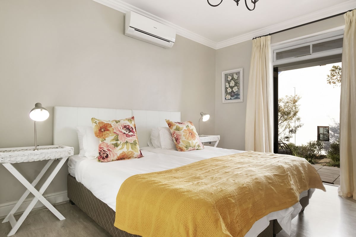 A bedroom with a bed, white nightstands, a white side table, a yellow throw blanket, and a window with curtains.