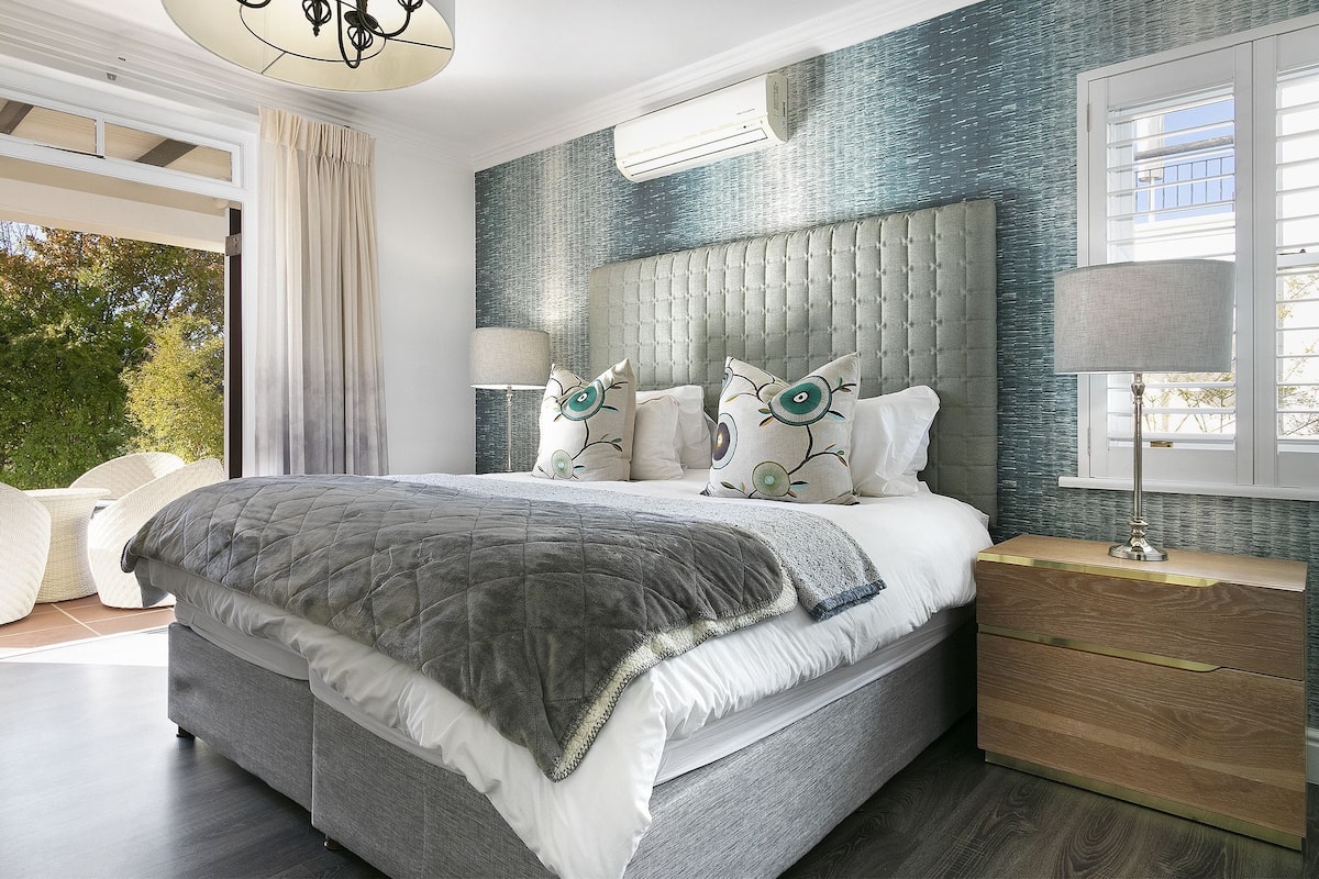 A modern bedroom with a large bed, a nightstand, and a view of greenery outside.