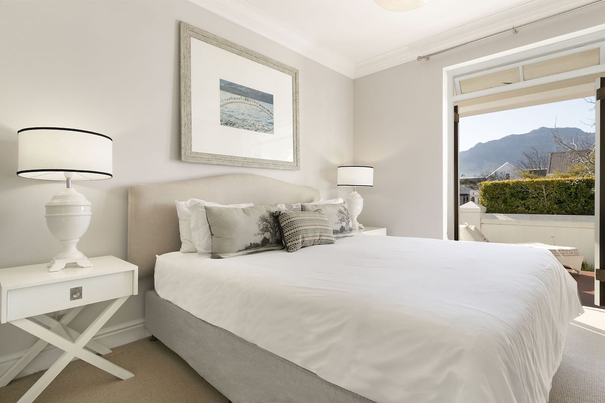A bedroom with a large bed, two bedside tables with lamps, a framed picture on the wall, and a view of mountains through an open door.
