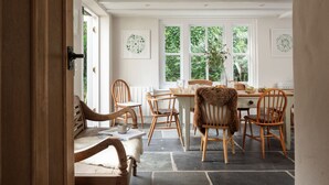 A dining area with wooden chairs and a table, large windows with greenery outside, and a white interior. - Calenick -  a stylish cottage that sleeps 8 guests in 4 bedrooms (Trelights)