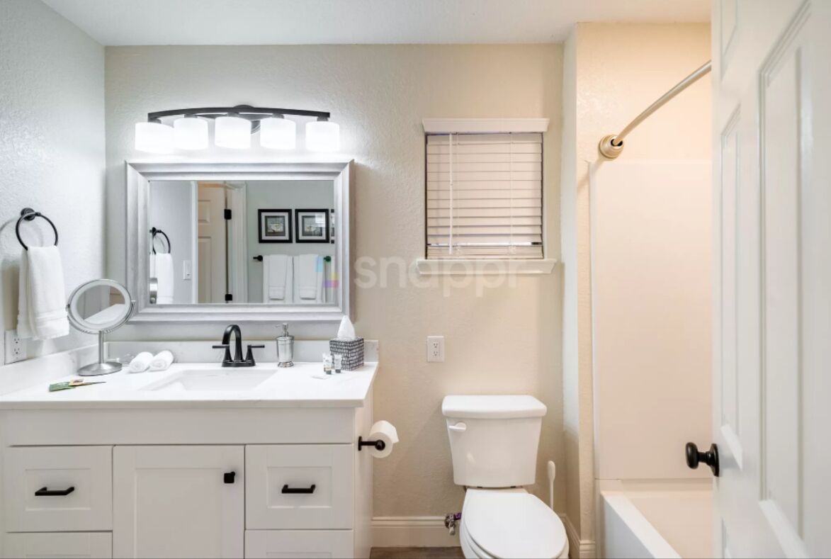 Un baño moderno con lavamanos, espejo y retrete blancos. Se observa una ventana con persianas y una zona de ducha.