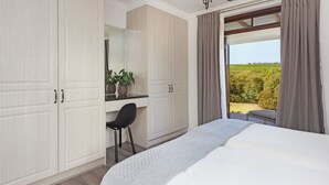 A bedroom with a large bed, a desk with a chair, a potted plant, and a view of a vineyard through a window. - Winelands Golf Lodges 1 (Cape Town)