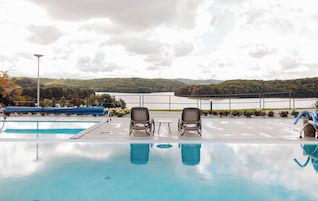 A swimming pool with two lounge chairs and a table, surrounded by a railing, with a view of a lake and forested hills.