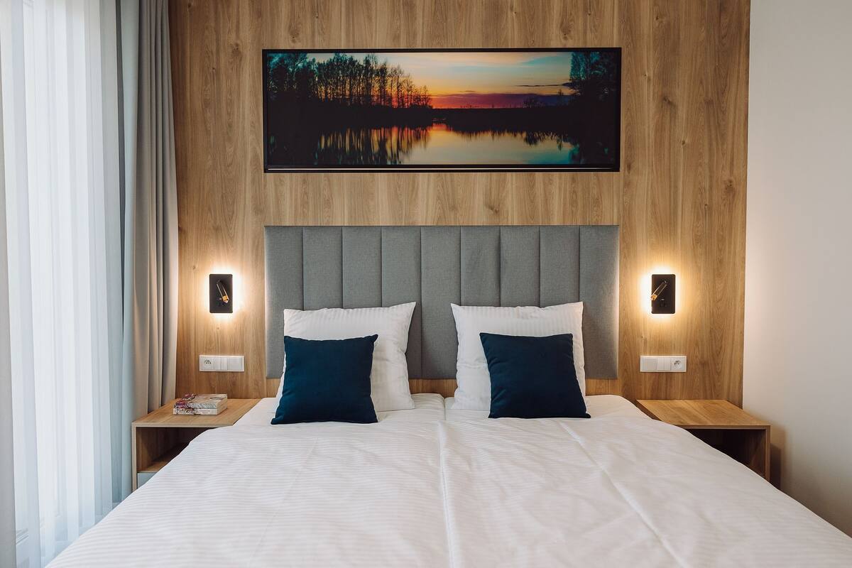 A double bed with a grey upholstered headboard, two bedside tables with wall-mounted lamps, and a framed landscape painting above the bed.