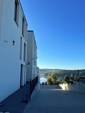 Una calle urbana estrecha con un edificio blanco a la izquierda, un cielo azul despejado y una vista distante de un lago y colinas.