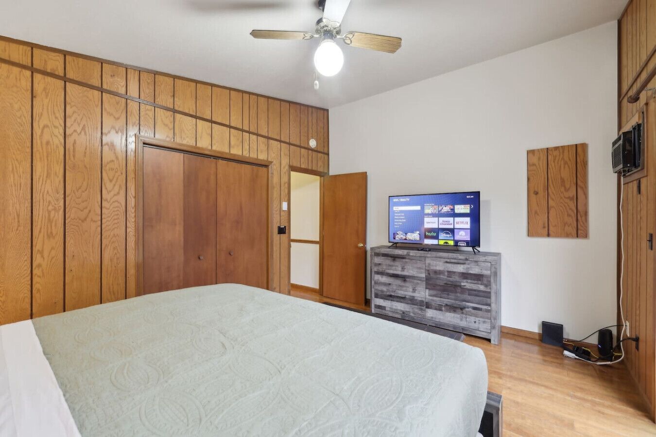 Un dormitorio con cama, armario de madera, televisor sobre un mueble y ventilador de techo.