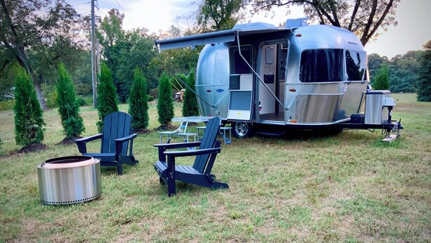 A recreational vehicle with a covered living area, two Adirondack chairs, and a portable grill. - Iconic Airstream River Glamping (Belton)
