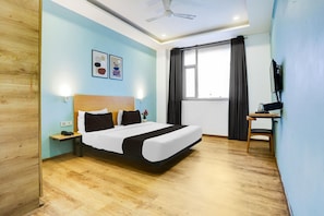 Una habitación de hotel con cama, escritorio, silla, ventilador de techo, ventana con cortinas y televisor montado en la pared.