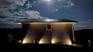 A single-story house with a sloped roof and a small window, illuminated by exterior lighting against a night sky with clouds and a visible moon. - A peaceful country retreat minutes from Watkins Glen—perfect for couples, friends, or family escapes. (Montour Falls)