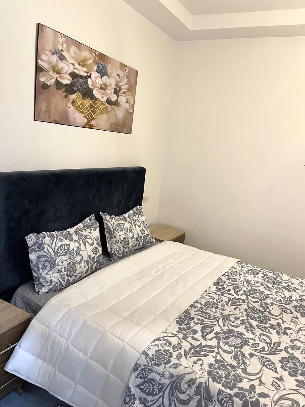 A bedroom with a dark headboard, a white quilted bedspread, and a floral painting on the wall.