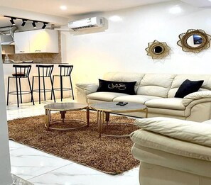 A modern living room with a beige sofa, a coffee table, and a dining area with chairs. - Cozy 1BR Retreat, Steps to Sand (Sousse)