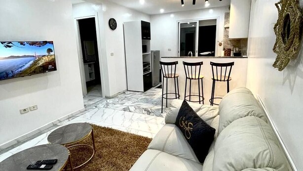 A modern living room with a white sofa, a coffee table, and a TV on the wall. There is a kitchen area in the background with a dining table and chairs. - Cozy 1BR Retreat, Steps to Sand (Sousse)