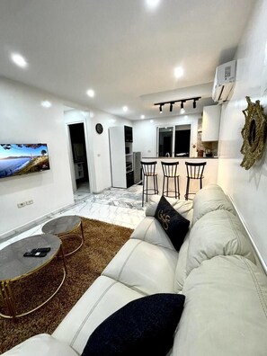 Una sala de estar moderna con un sofá blanco, una mesa de centro y un televisor en la pared. Se observa una zona de cocina al fondo con una mesa y sillas.