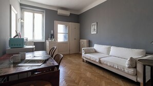 A room with a wooden herringbone floor, a white sofa, a dining table with chairs, a window with curtains, and a radiator. - Italianway Easy - Rosmini 13 B (Milan)