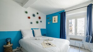 Een net opgemaakt bed met witte lakens, een blauwe accentmuur en een raam met blauwe gordijnen.