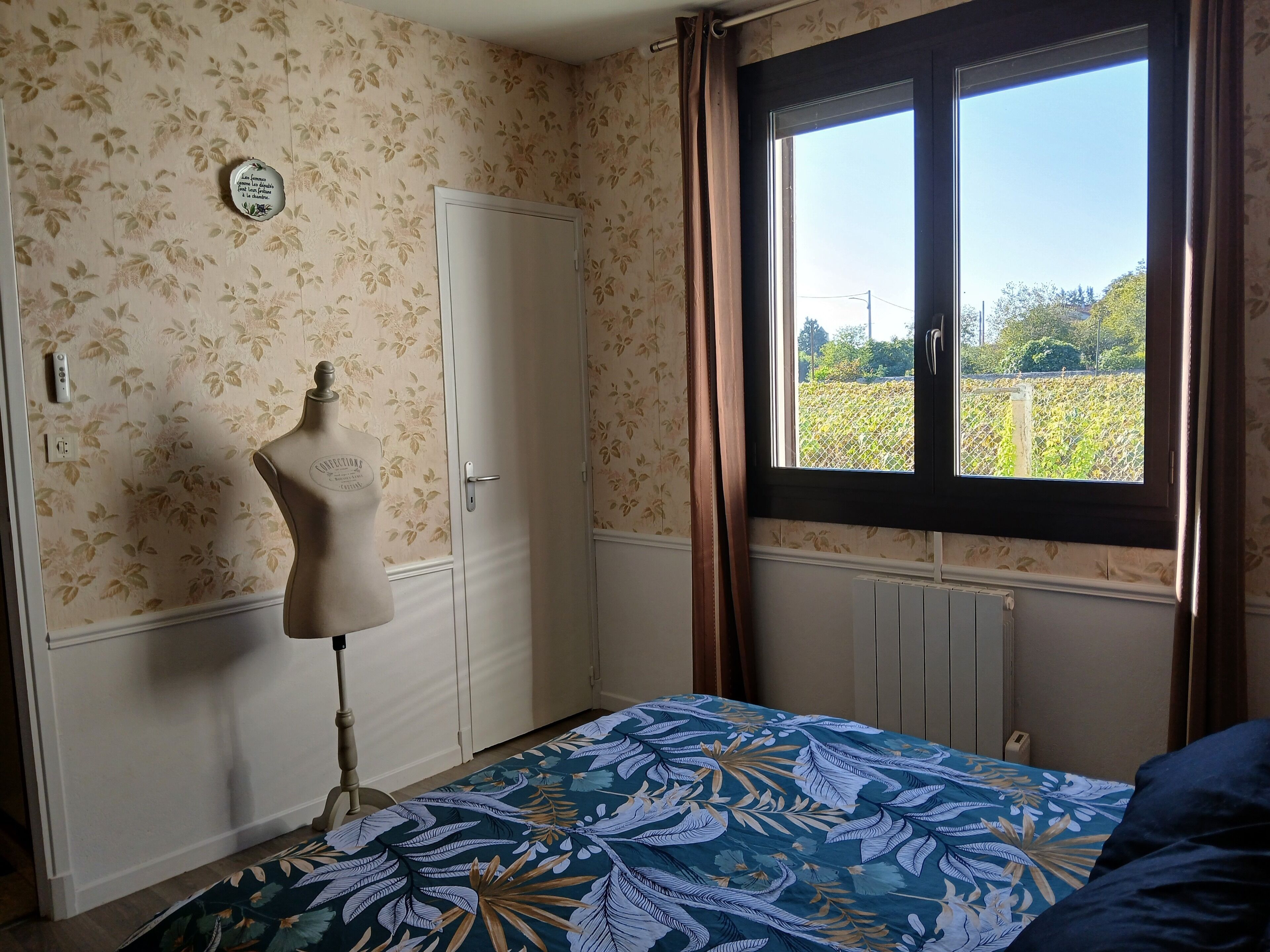 Ein Zimmer mit floralem Wandpapier, einem weißen Kleiderschrank, einer Mannequin, einem Fenster mit Blick ins Grüne und einem Bett mit einer blumigen Tagesdecke.