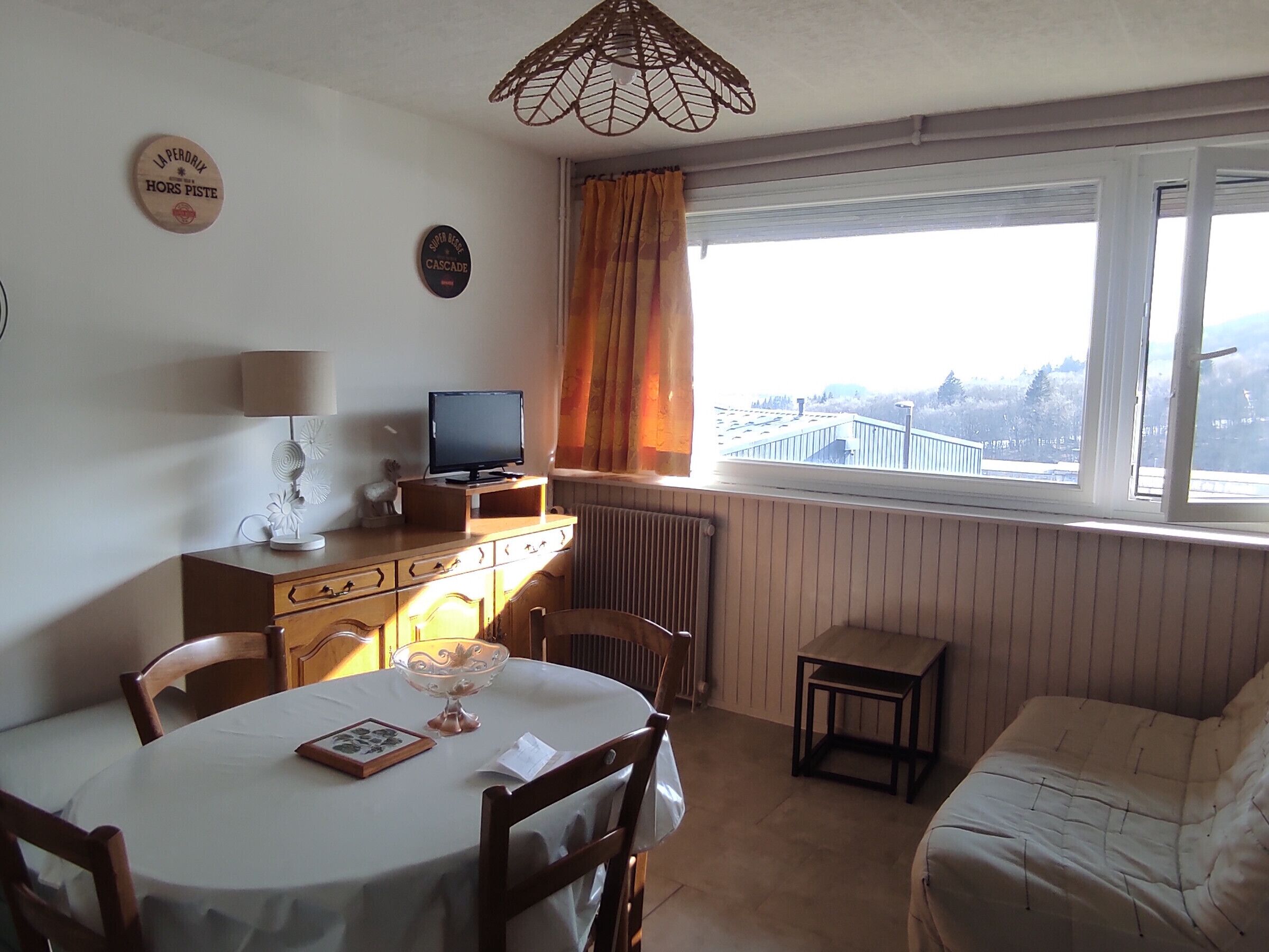 Una habitación con una mesa de comedor, sillas, un televisor sobre un mueble de madera, un radiador, una ventana con cortinas y vistas a un puente.