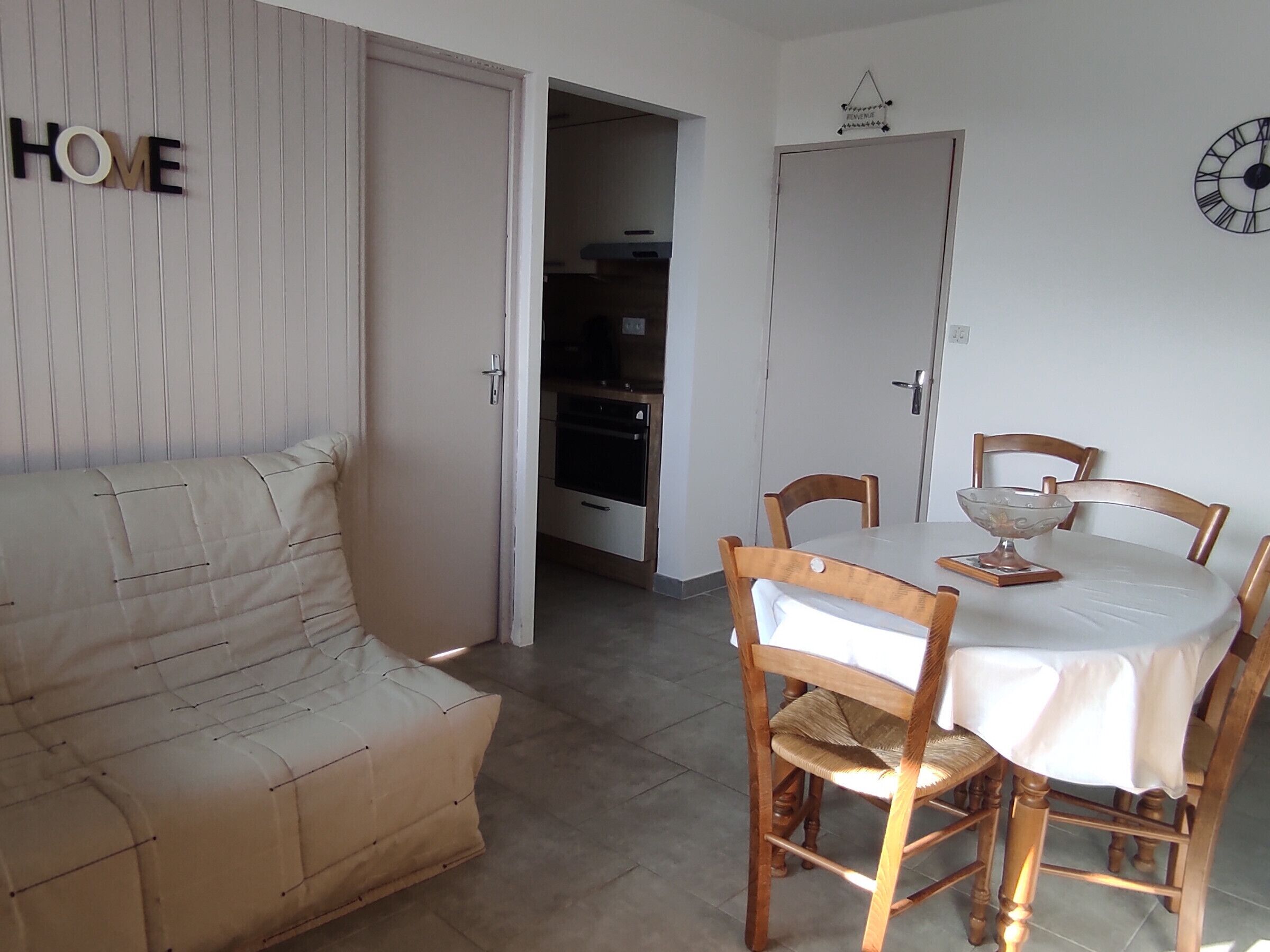 A living room with a sofa, a dining table with chairs, a clock on the wall, and a door leading to another room.