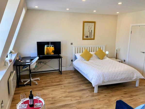 A bedroom with a bed, a desk with a laptop, a television, and a dining table with wine glasses and a bottle.